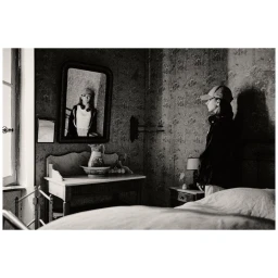 Black and white portrait of a woman in profile wearing a cap, standing in a bedroom with patterned wallpaper, mirror, and washstand.