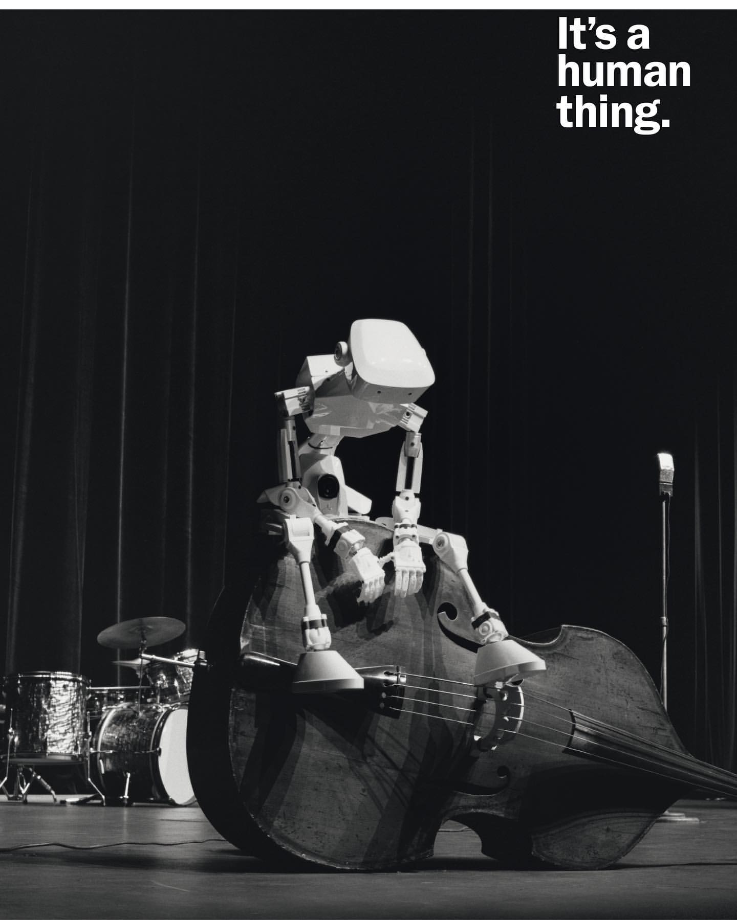 Robot sitting on a contrabass on a stage, grayscale image. Drums and other musical instruments in the background, vintage microphone stand.