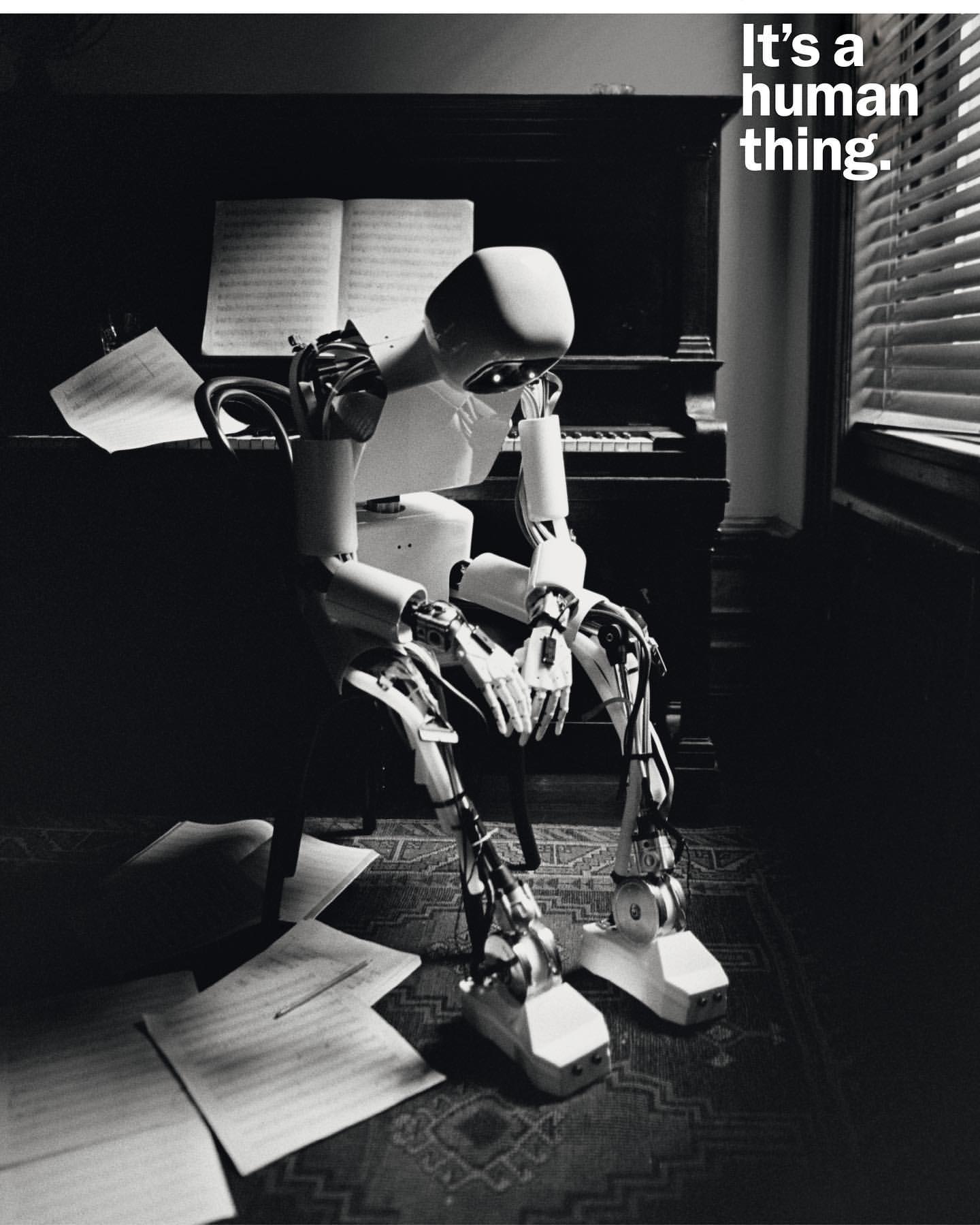 Monochrome image of a humanoid robot seated at a piano with scattered sheet music, expressing a contemplative mood in a dimly lit room.