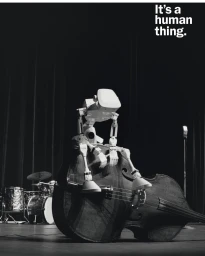 White robotic figure seated on its side atop a large double bass on a dark stage with a drum kit in the background