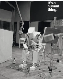 A white humanoid robot seated on a folding chair in a recording studio with a trumpet on the floor, photographed by Vincent Dixon.