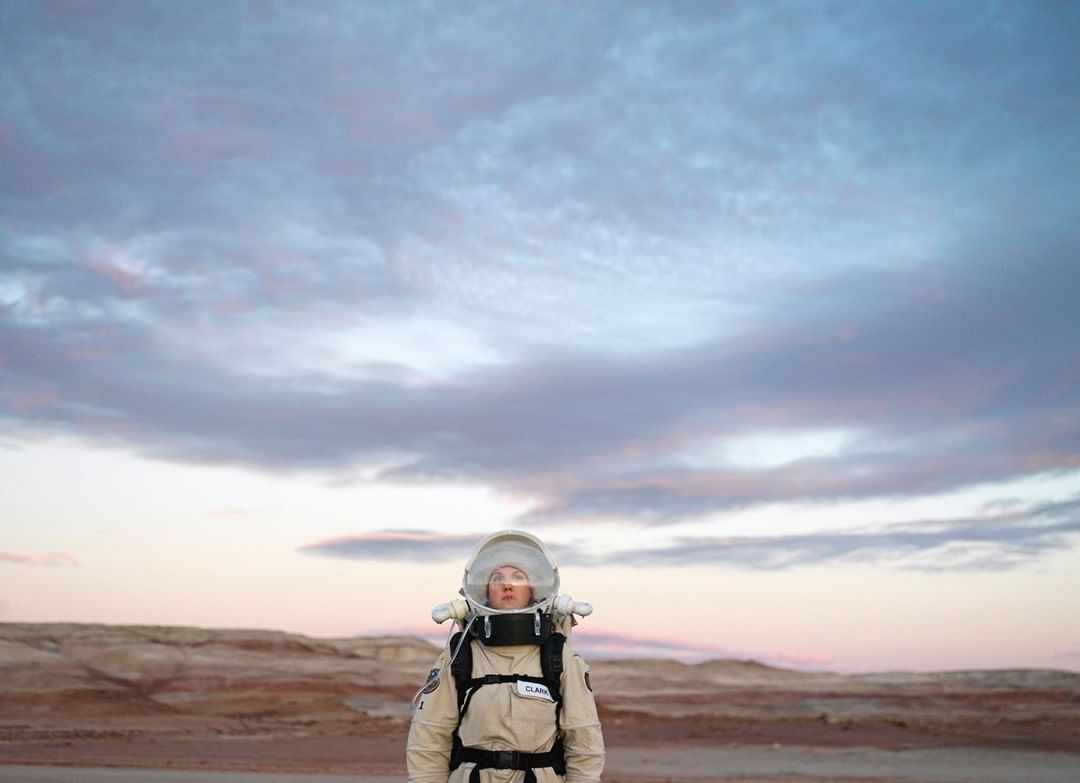 Astronaut in a space suit stands on a red, rocky landscape against a sky with pink and purple clouds at sunrise or sunset.