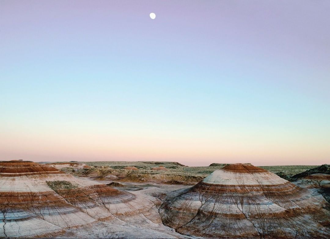 Scenic view of layered rock formations in a desert landscape under a pastel sky with a visible moon, captured at dusk.