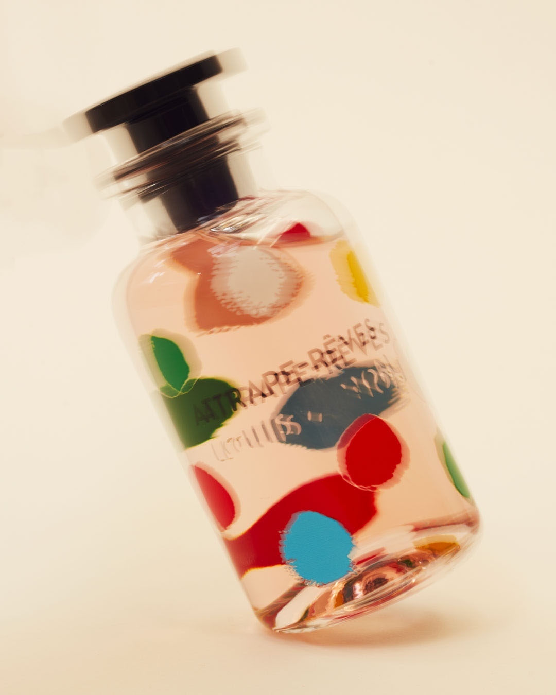 Close-up studio shot of a perfume bottle with a polka dot design, featuring pink liquid, a black cap, and the label 'Attrape-Rêves'.