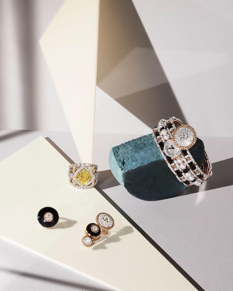 High-angle shot of gold and gemstone jewelry, including a bracelet, rings, and earrings, arranged against a modern geometric backdrop.