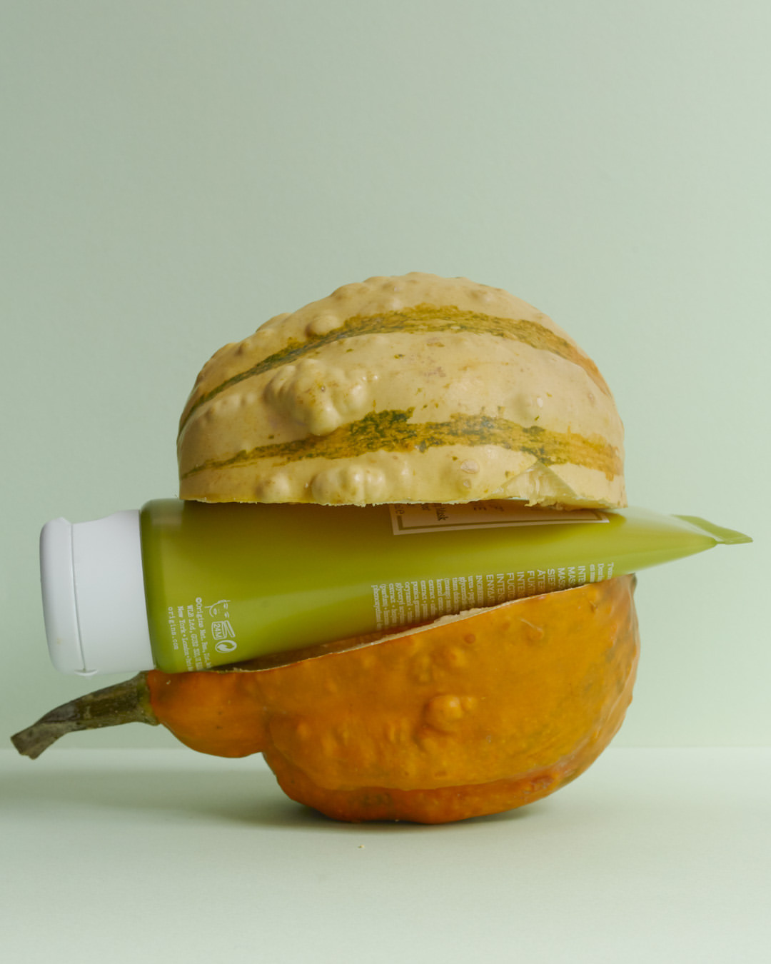Still life of green cosmetic tube nestled between two halves of an orange and cream striped gourd against light green background.