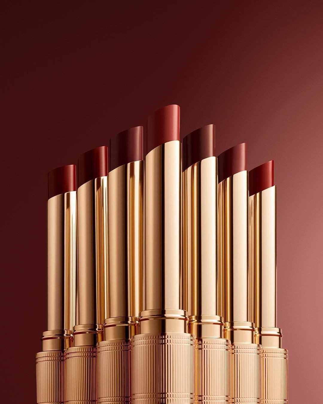 Seven shades of lipstick in a row with gold cases. The lipstick colors range from red to maroon, displayed on a gradient background.
