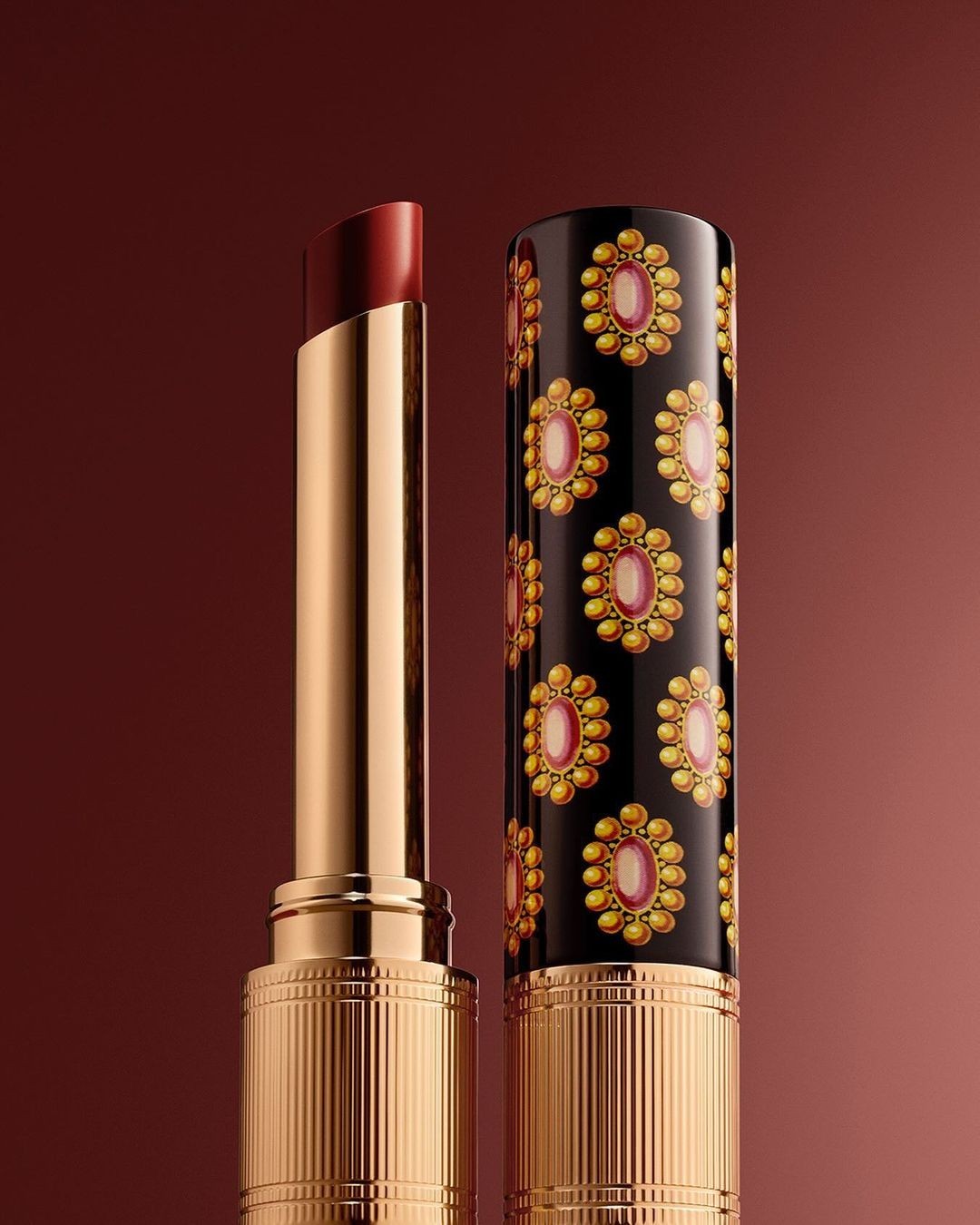 Close-up of a dark red lipstick, partially extended from its gold and black patterned tube, against a graduated brown background.