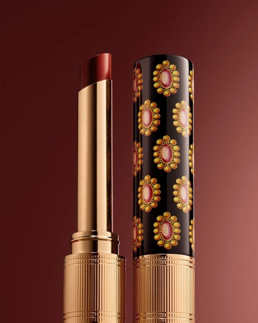 Close-up of a dark red lipstick, partially extended from its gold and black patterned tube, against a graduated brown background.