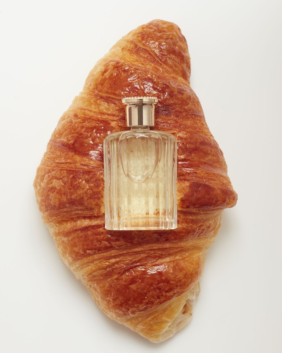 Close-up studio shot of a croissant with a glass perfume bottle on top. The scene is set against a white background.