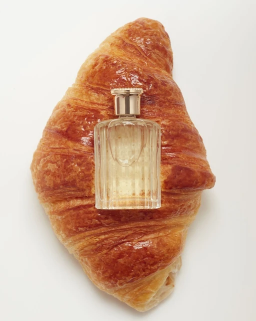 Close-up studio shot of a croissant with a glass perfume bottle on top. The scene is set against a white background.