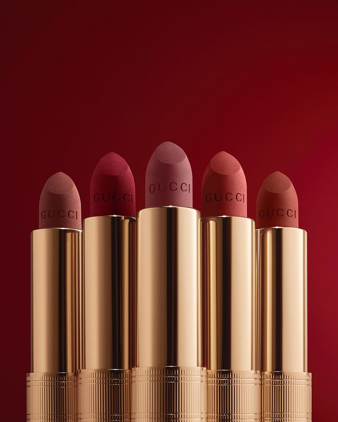 Close-up studio shot of five golden Gucci lipstick tubes with matte lipstick of varying brown and red shades against a red background.