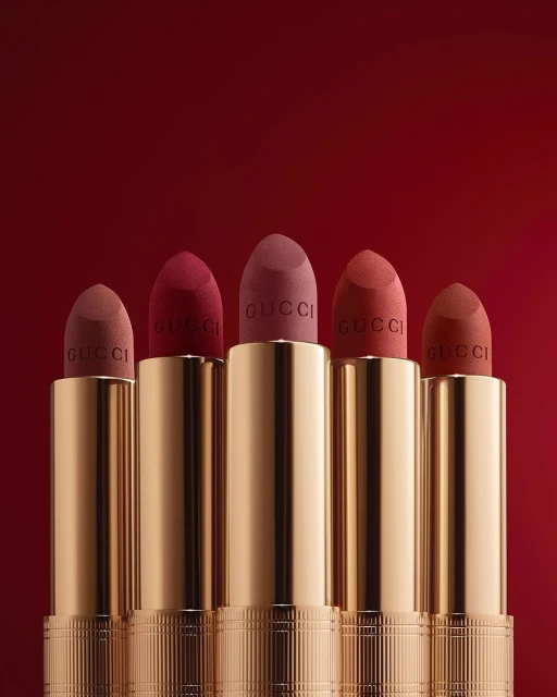 Close-up studio shot of five golden Gucci lipstick tubes with matte lipstick of varying brown and red shades against a red background.