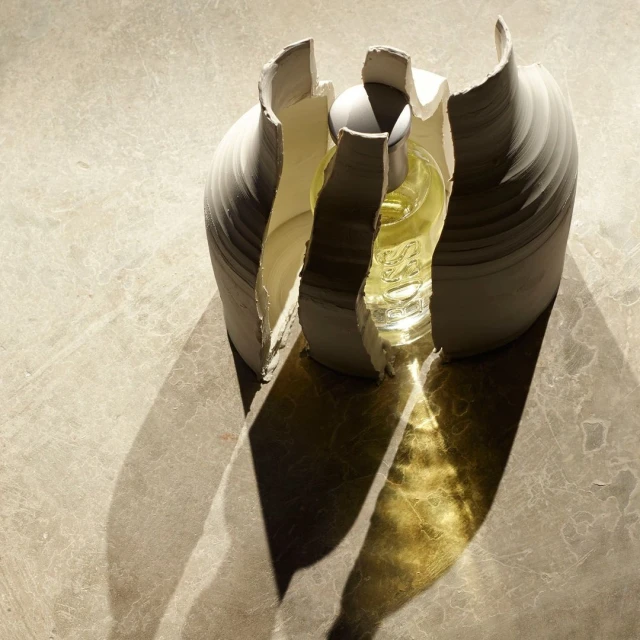 Glass bottle with yellow liquid inside a decorative white ceramic vase, set on a neutral surface with defined shadows.