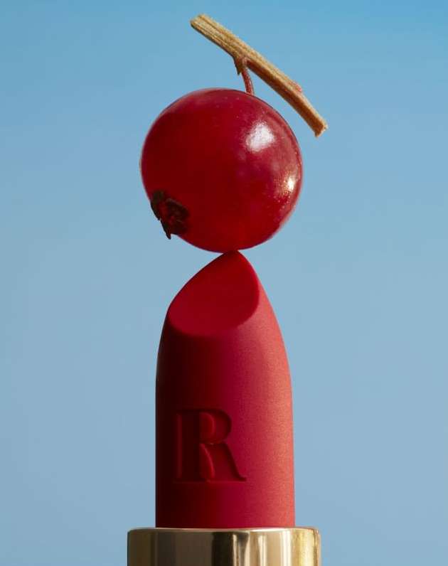 Close-up studio shot of a red lipstick with a red berry on top against a blue background, beauty and cosmetics product shot.