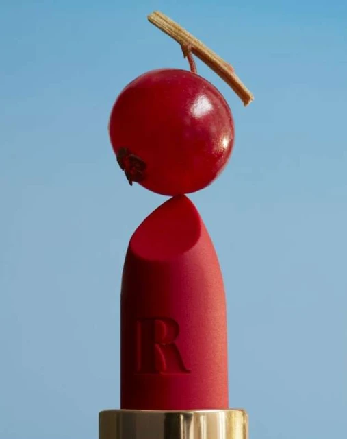 Close-up studio shot of a red lipstick with a red berry on top against a blue background, beauty and cosmetics product shot.