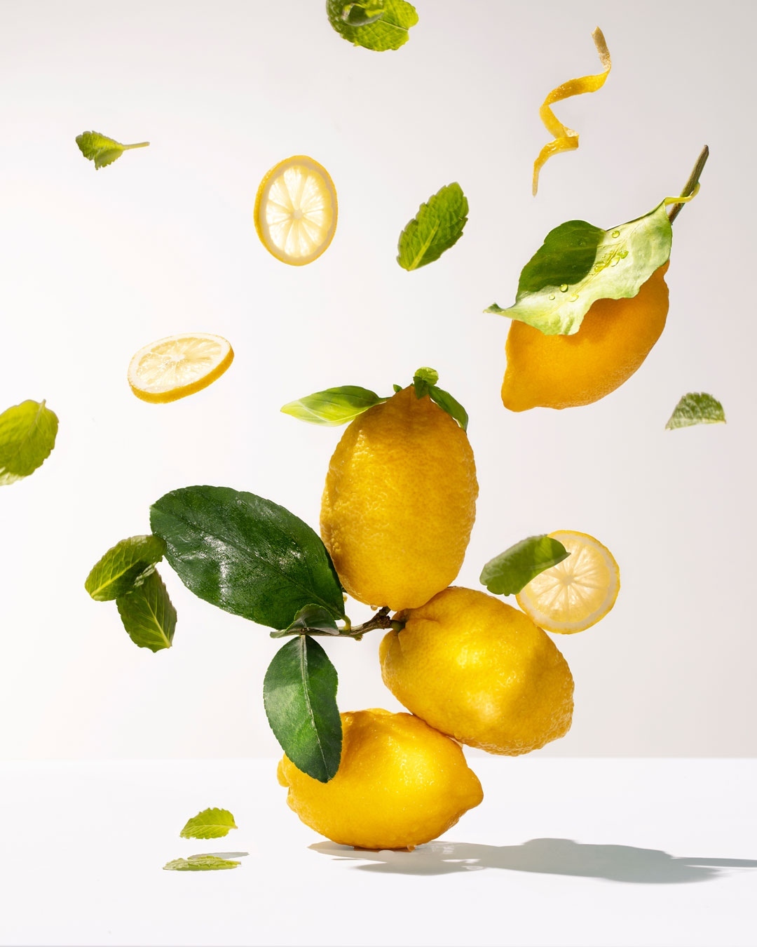 Still life of lemons stacked with fresh mint leaves, lemon slices, and lemon peel floating in the air against a white background.