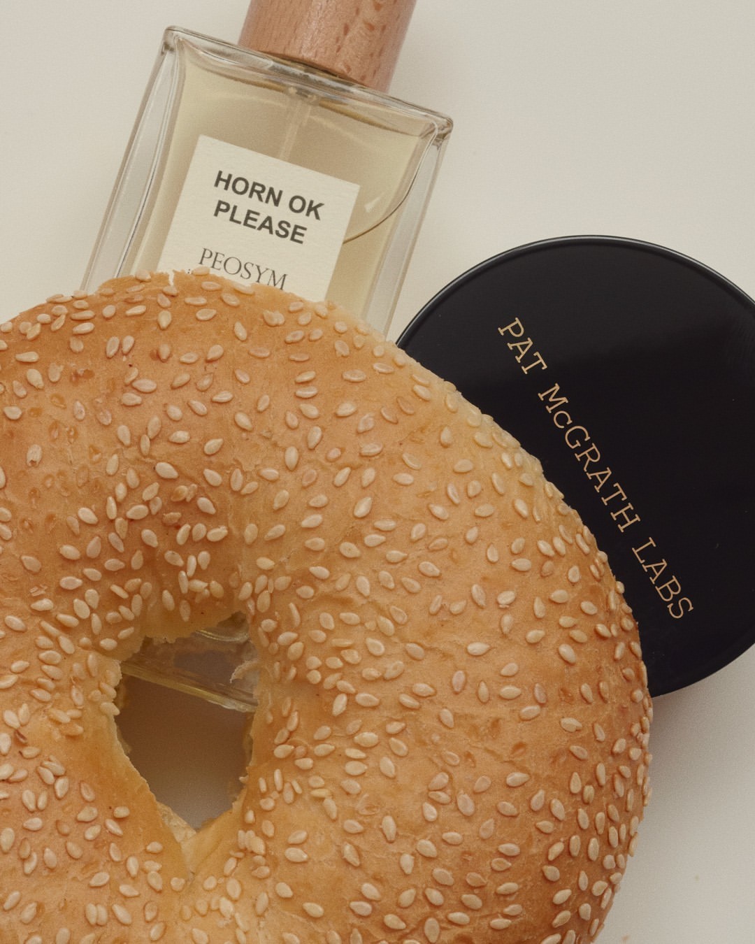 Close-up of a sesame bagel next to a bottle of Horn OK Please perfume and Pat McGrath Labs compact on a white surface.