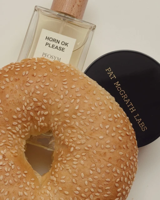 Close-up of a sesame bagel next to a bottle of Horn OK Please perfume and Pat McGrath Labs compact on a white surface.