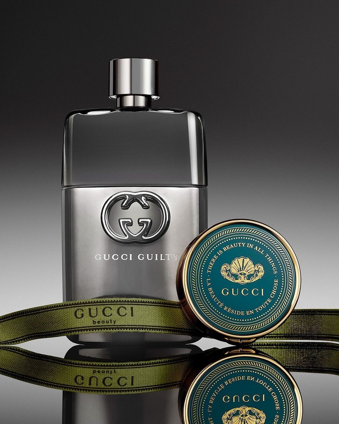 Gucci Guilty perfume bottle on a reflective surface with a Gucci beauty compact, set against a gradient gray backdrop in a studio setting.