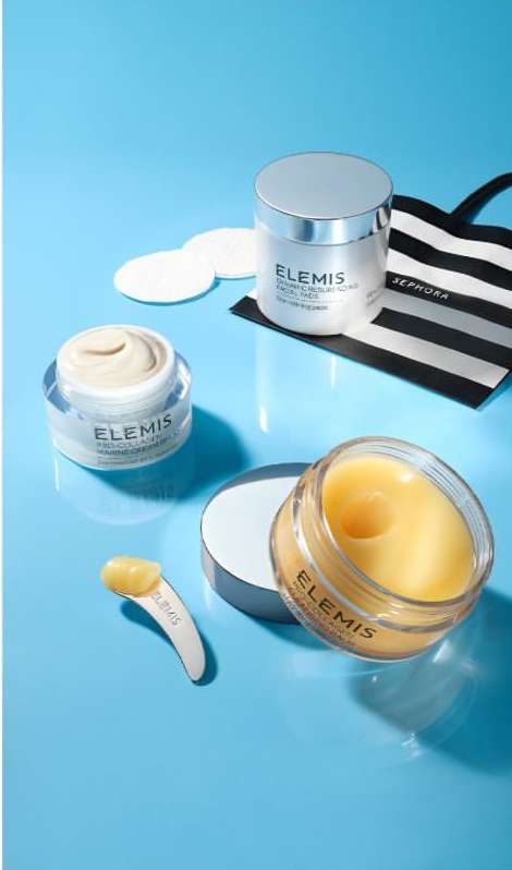Elevated shot of three Elemis skin care products, cotton pads, and a striped Sephora bag on a bright blue surface, product-focused.