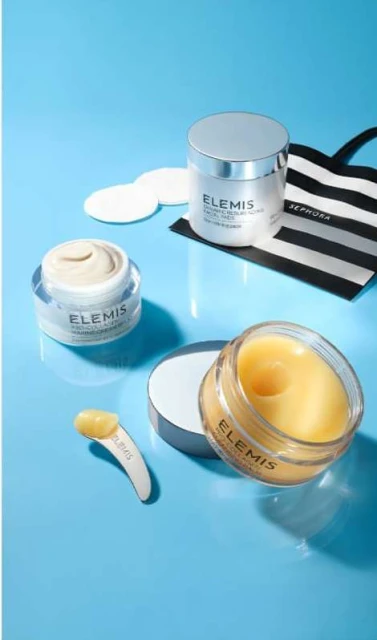 Elevated shot of three Elemis skin care products, cotton pads, and a striped Sephora bag on a bright blue surface, product-focused.