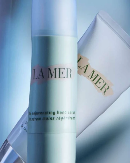 La Mer skincare products feature a bottle of rejuvenating hand serum and a tube against a light blue gradient background in a studio shot.
