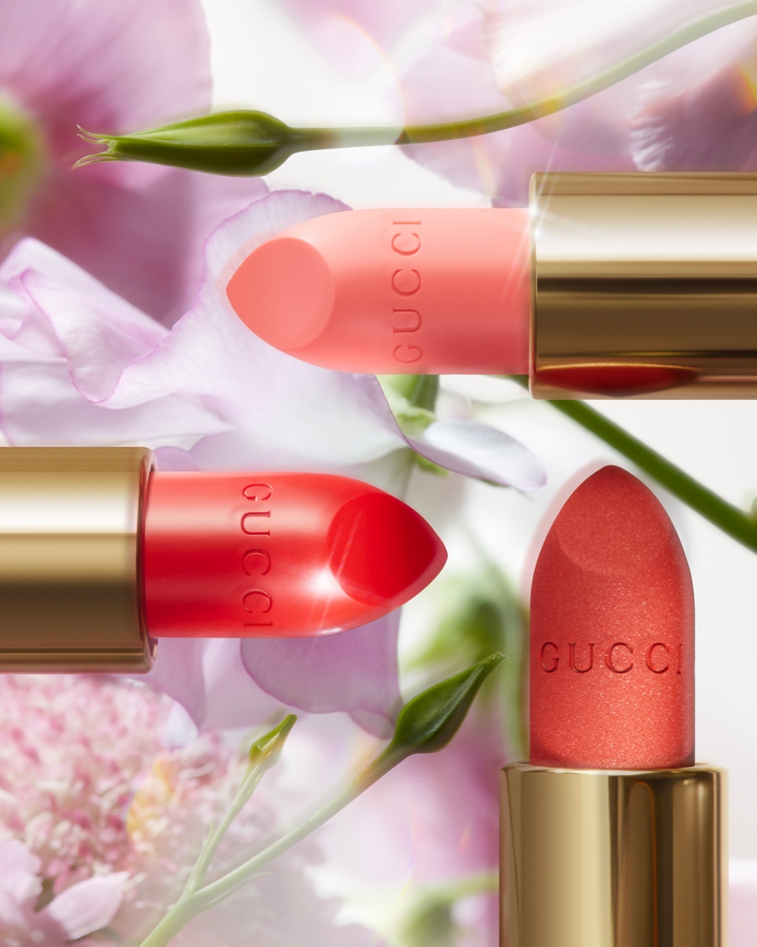 Three Gucci lipsticks in coral, red, and orange with gold tubes surrounded by a soft-focus floral background create a beauty product shot.
