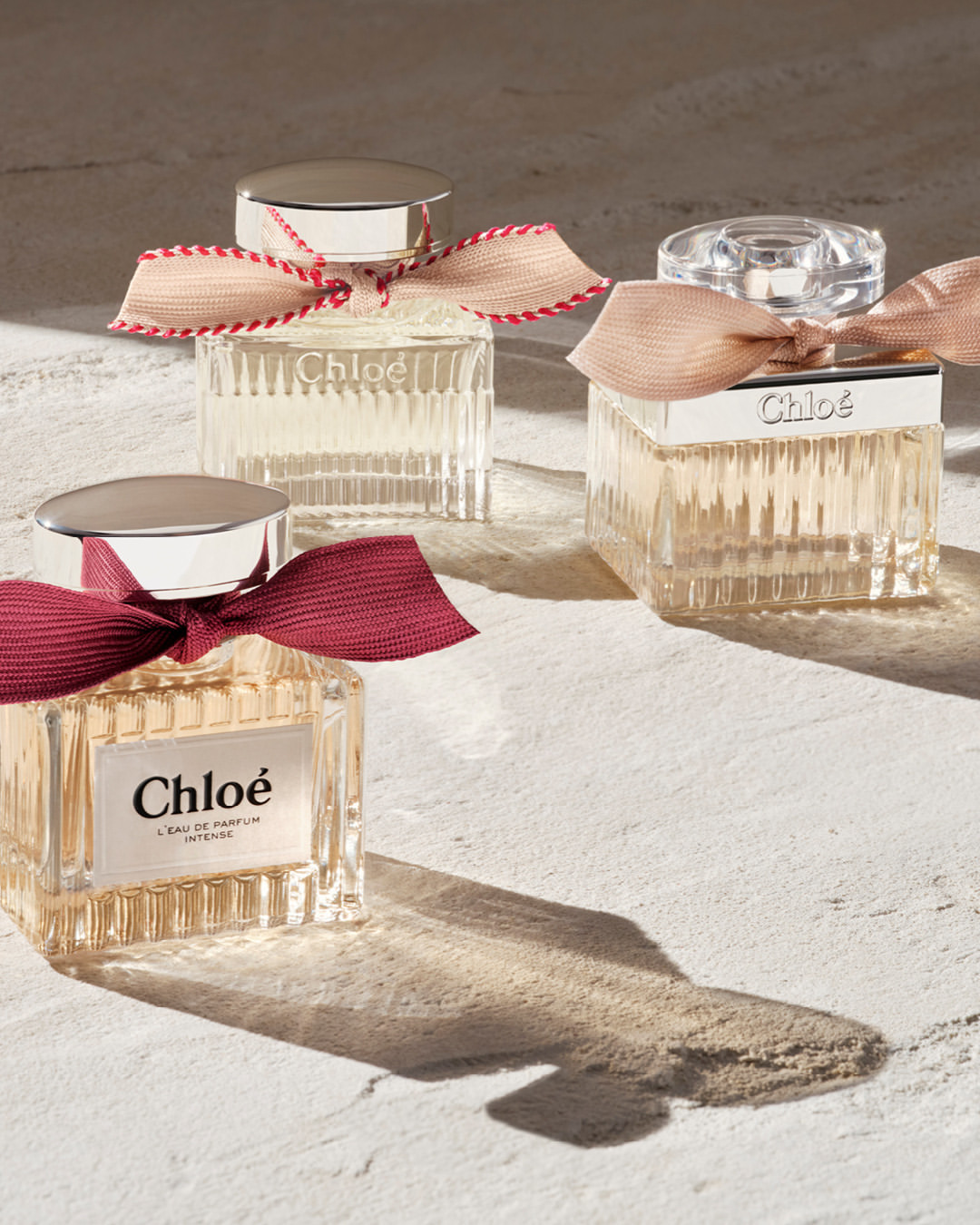 Three Chloe perfume bottles with decorative ribbons on a textured beige surface, showcasing a high-end beauty product presentation.