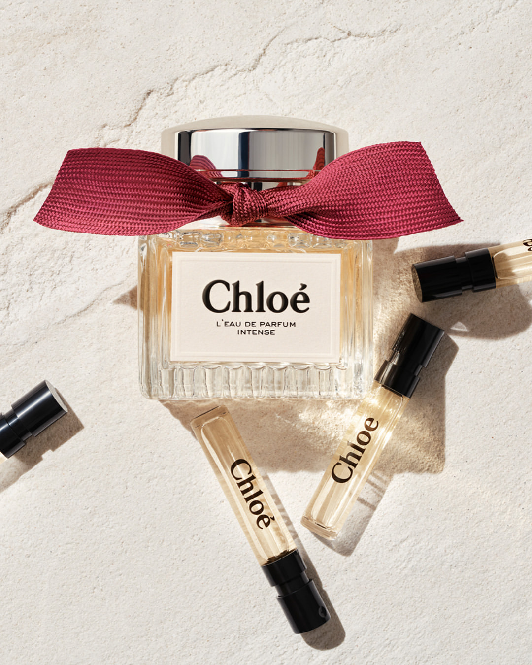Chloe perfume bottle with red ribbon, surrounded by sample vials, all on a textured white surface in soft, diffused lighting.