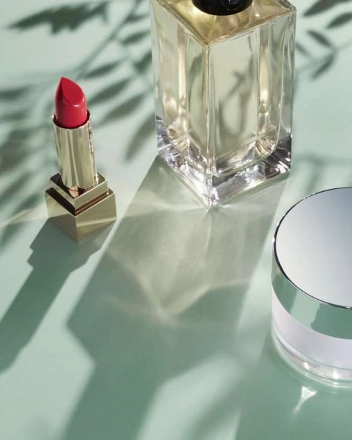 Beauty product photography: lipstick tube, fragrance bottle, and cosmetic container on a light green surface with leaf shadows.