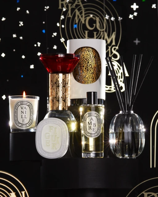 Luxury candles and fragrance products on a black platform against a dark backdrop featuring gold and white graphic accents, studio lighting.