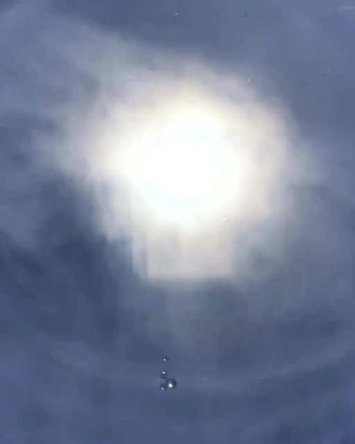 An abstract view of the sun with dark clouds and a few raindrops, creating a dreamy, atmospheric effect. The light is bright and diffused.
