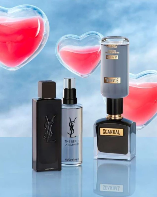 Close-up shot of Yves Saint Laurent and Scandal perfume bottles with heart-shaped transparent decor against a light blue background.