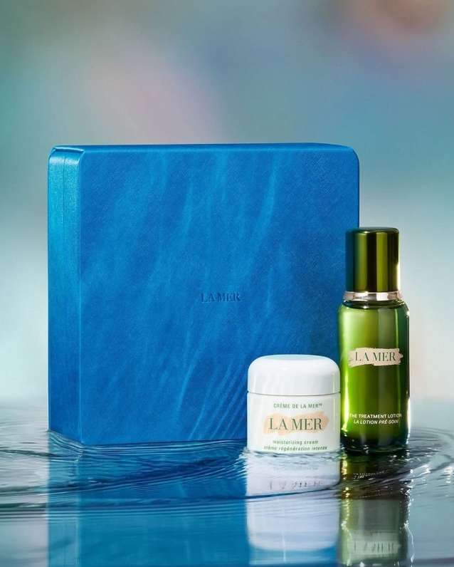 La Mer skincare set featuring a blue box, cream jar, and green bottle on a reflective watery surface against a blurred background.