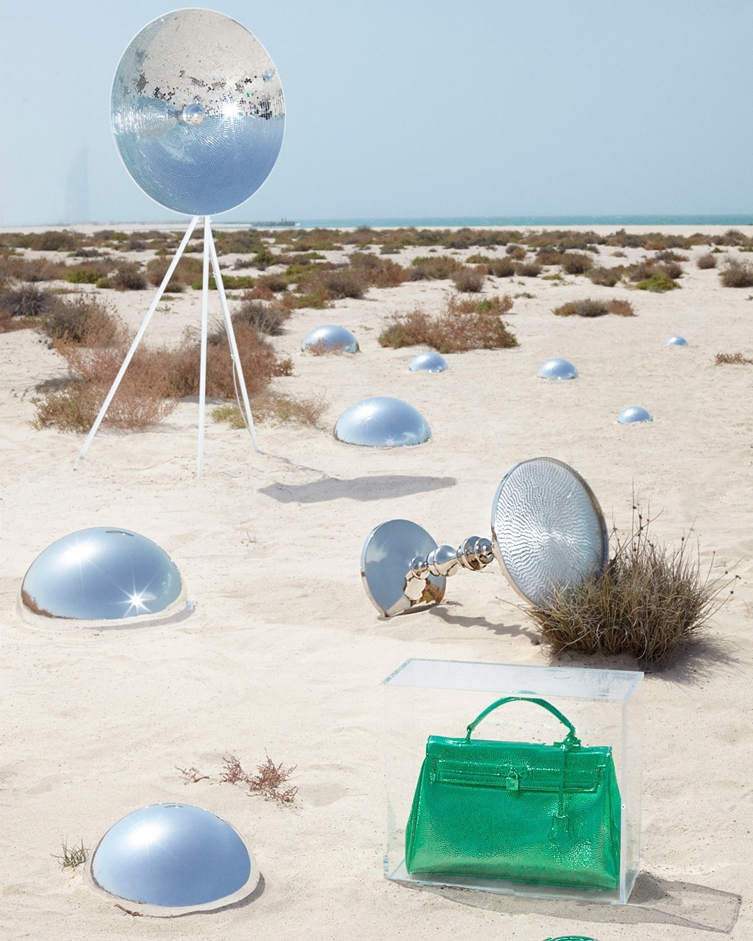 Fashion still life in a desert featuring a bright green handbag in a clear acrylic box, set among reflective dome objects and tripods.