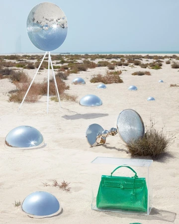 Fashion still life in a desert featuring a bright green handbag in a clear acrylic box, set among reflective dome objects and tripods.