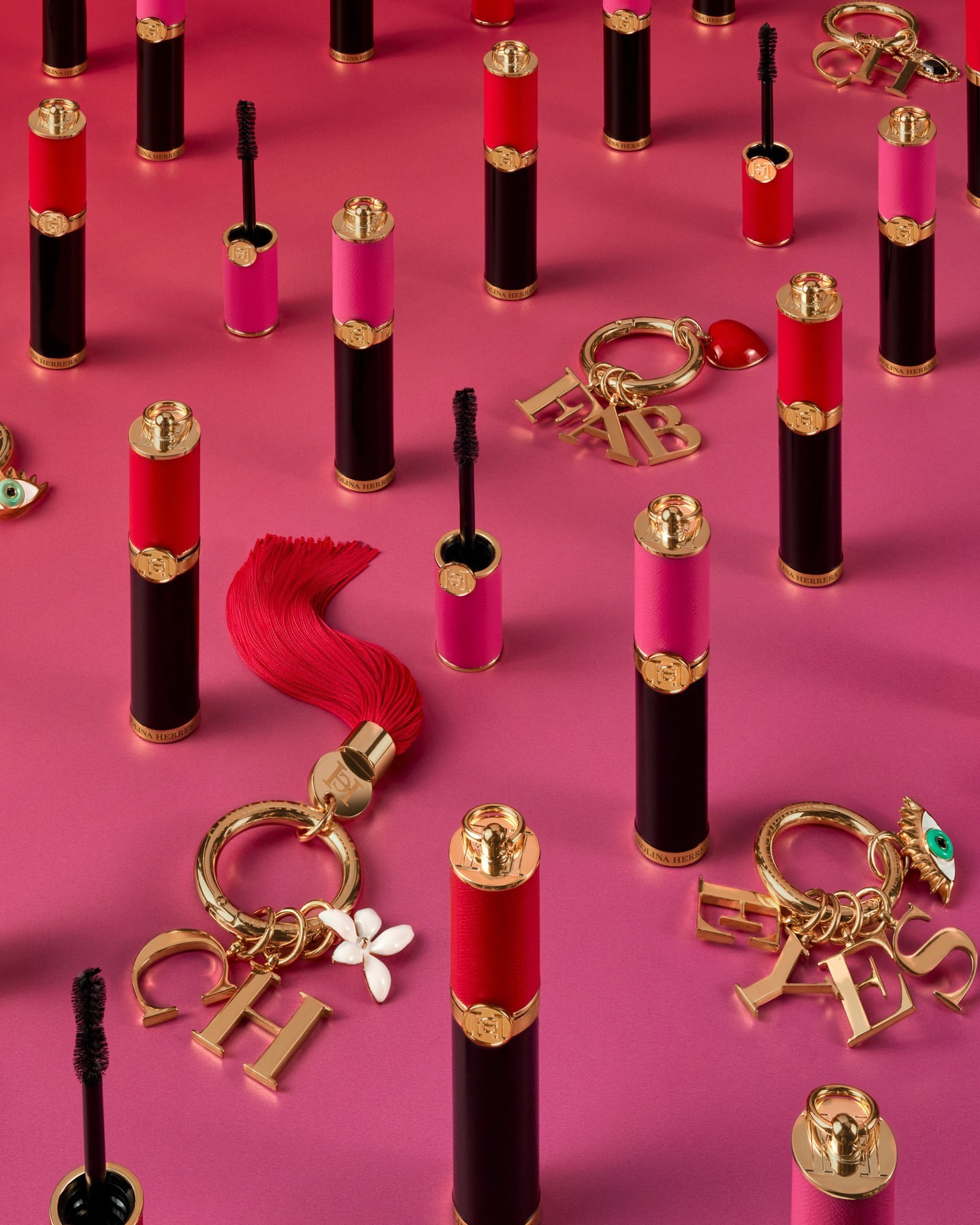 Eye-level shot of black, red, and pink mascara tubes arranged on a pink surface, interspersed with golden keychains and letter charms.