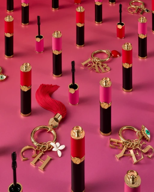 Eye-level shot of black, red, and pink mascara tubes arranged on a pink surface, interspersed with golden keychains and letter charms.