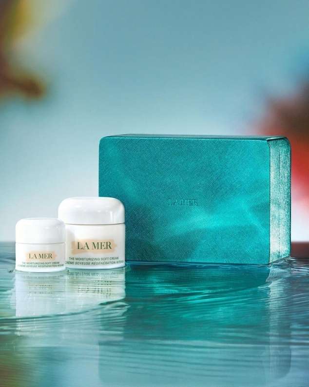 La Mer skincare set with two jars of moisturizing cream and a gift box on a reflective surface with a light blue background.