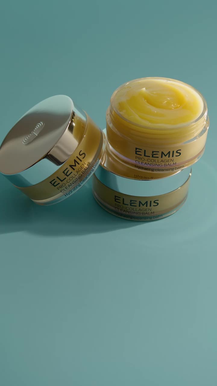 Three Elemis Pro-Collagen Cleansing Balm jars stacked on a blue surface. The jars feature silver lids and minimalist label design.