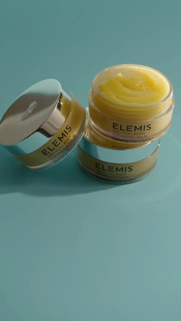 Three Elemis Pro-Collagen Cleansing Balm jars stacked on a blue surface. The jars feature silver lids and minimalist label design.