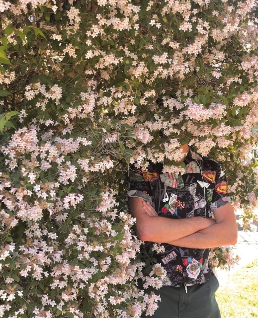 Person with arms crossed stands behind flowering bush outdoors wearing patterned short-sleeve shirt and dark green pants.