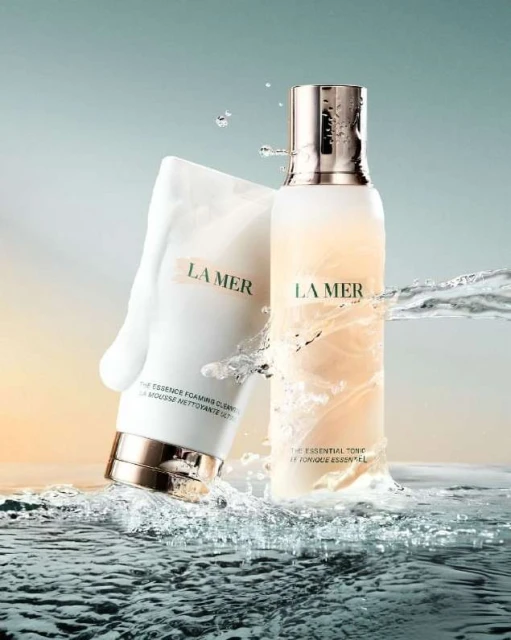 La Mer skincare products are displayed with water elements. A white tube and a bottle are featured, surrounded by water splashes.