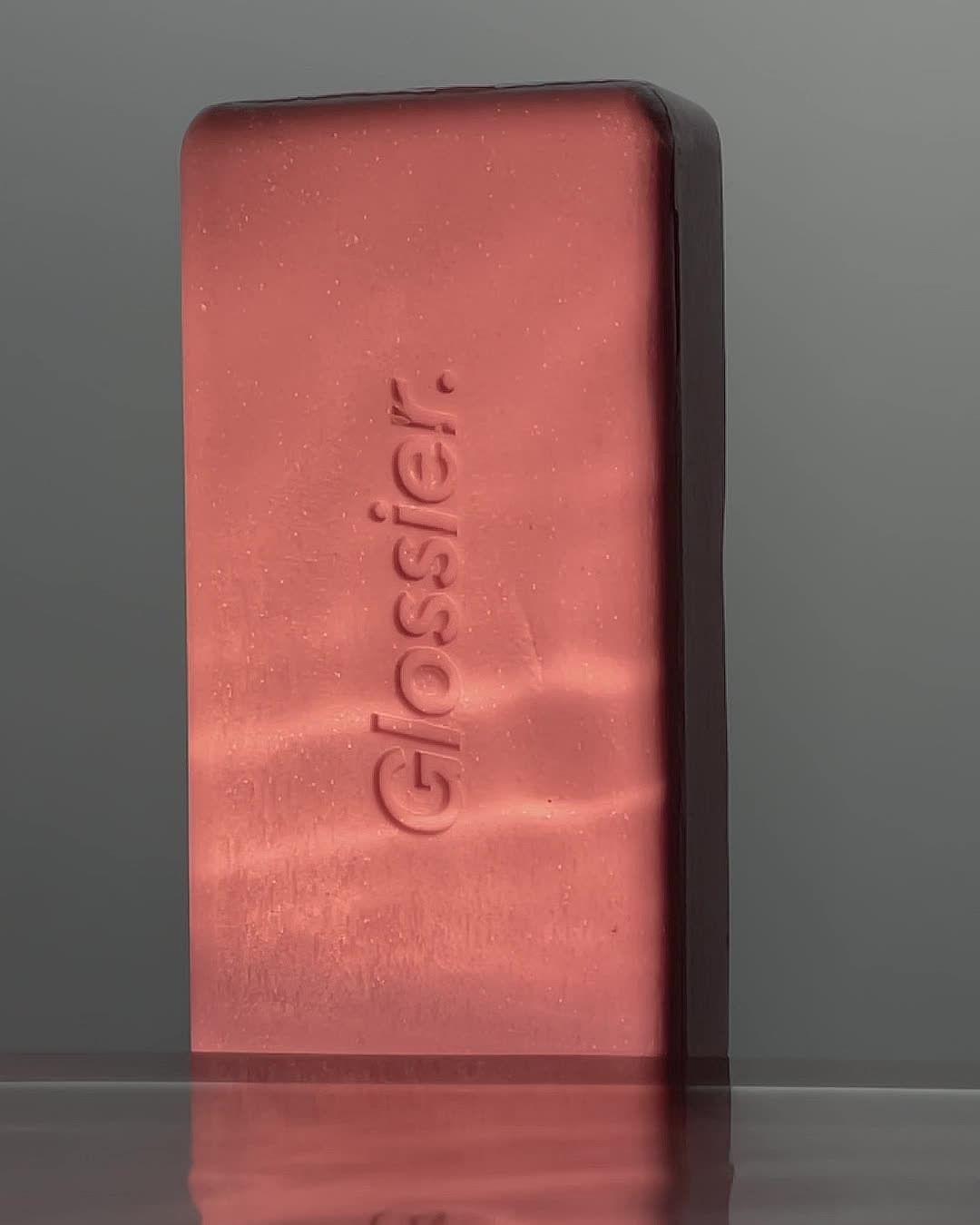 Close-up of Glossier bronzer compact in sleek, minimalist packaging, reflecting light on a glossy surface, studio shot.