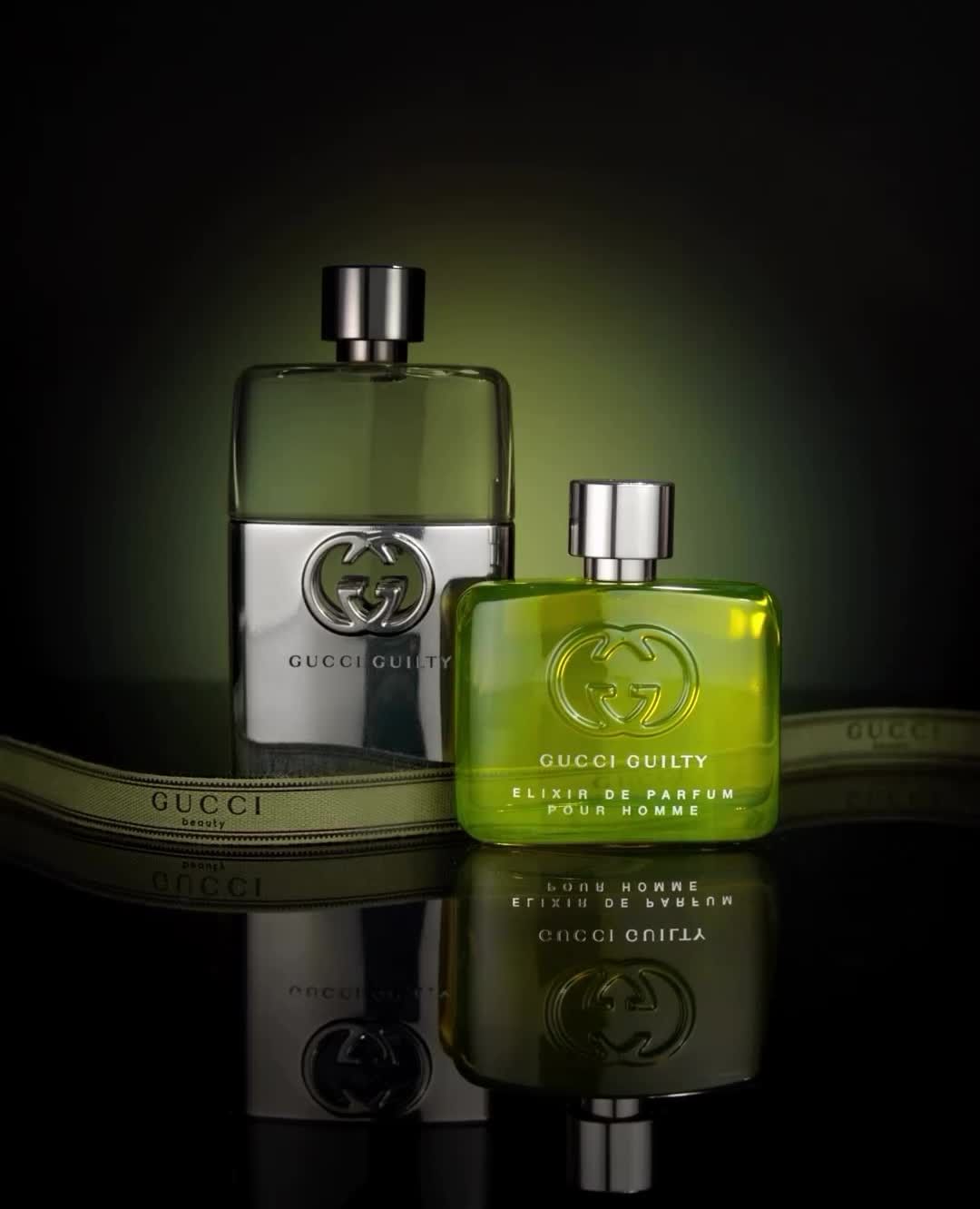 Two Gucci Guilty cologne bottles with silver and green liquid on a reflective surface with an olive green ribbon detail against a dark background.