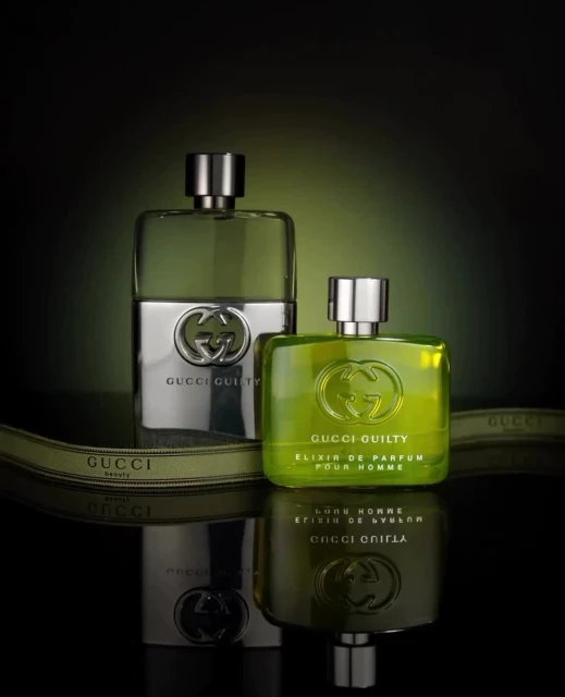 Two Gucci Guilty cologne bottles with silver and green liquid on a reflective surface with an olive green ribbon detail against a dark background.