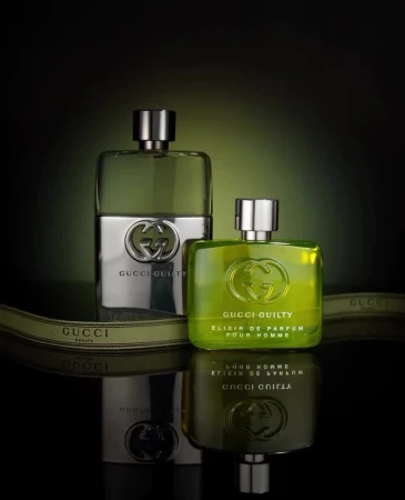 Two Gucci Guilty cologne bottles with silver and green liquid on a reflective surface with an olive green ribbon detail against a dark background.