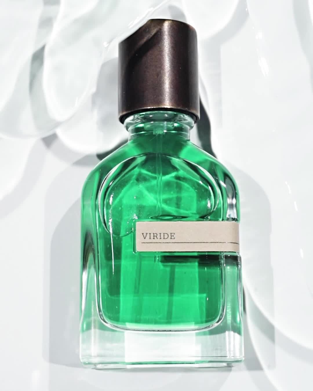 A green glass perfume bottle with a dark cap sits on a white surface with light reflecting, creating a clean and luxurious aesthetic.