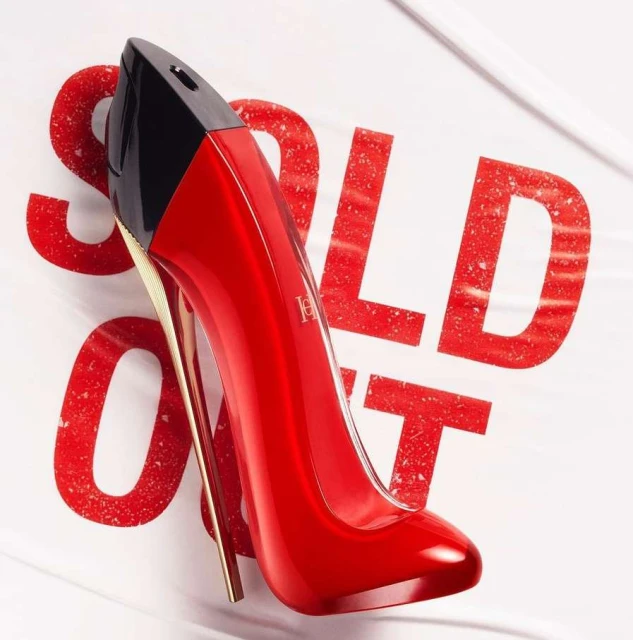 Shiny red stiletto-shaped Carolina Herrera Good Girl perfume bottle with gold heel and black top, against off-white textured background.